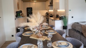 Un coin repas avec une longue table dressée pour six personnes, entourée de chaises à revêtement gris. Un vase contenant des plumes d’herbe des pampas et une petite plante en pot sont posés sur la table.