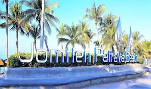 A sign with the text "Jomien Pattaya Beach" surrounded by palm trees and a clear sky.