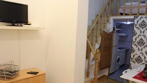 A room with a TV on a shelf, a wooden cabinet, a table with a remote, and a staircase leading to a laundry area. - Apartment in front of Plaza Mayor and Sol (Madrid)