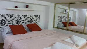 A neatly made bed with a patterned bedspread and two pillows, a large mirror on the wall, and a small shelf with a few items. - Apartment in front of Plaza Mayor and Sol (Madrid)