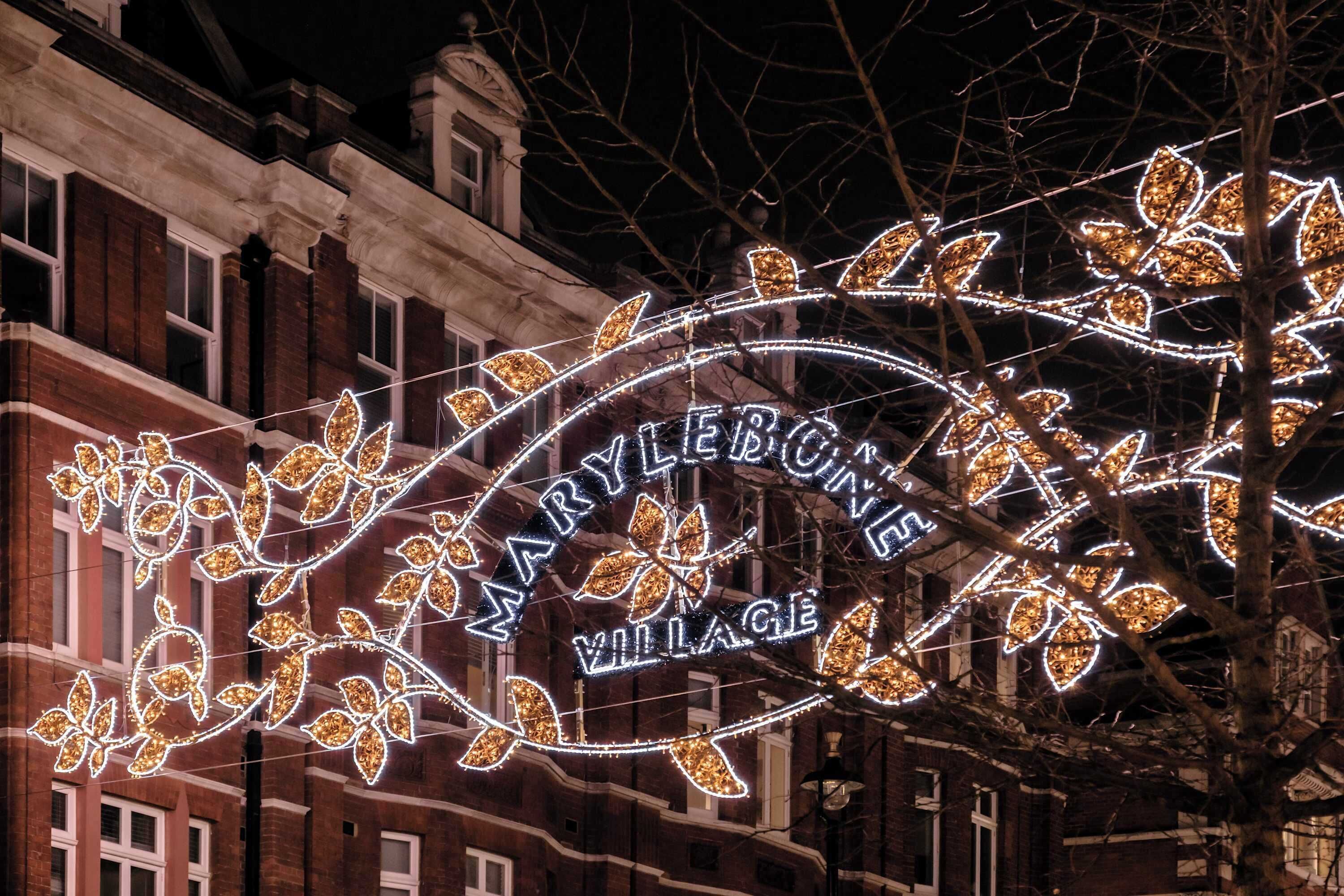 A sign with illuminated leaf designs and the text "MARYLEBONE VILLAGE" against a brick building backdrop.