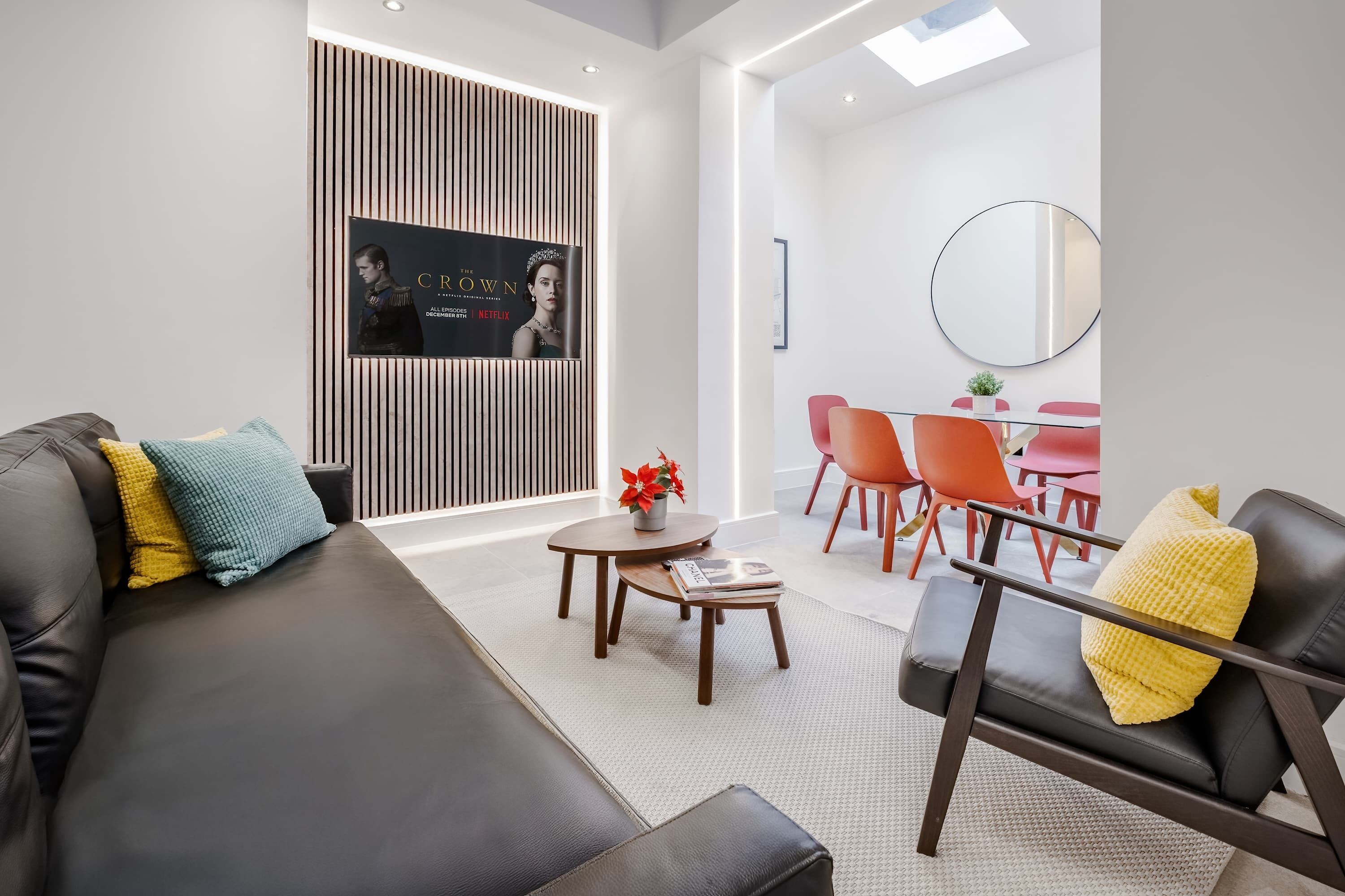 Un salon moderne avec un canapé en cuir noir, une petite table en bois sur laquelle repose un magazine, et un téléviseur fixé au mur, ceint de stores verticaux.