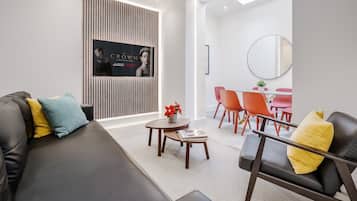 Un salon moderne avec un canapé en cuir noir, une petite table en bois sur laquelle repose un magazine, et un téléviseur fixé au mur, ceint de stores verticaux.