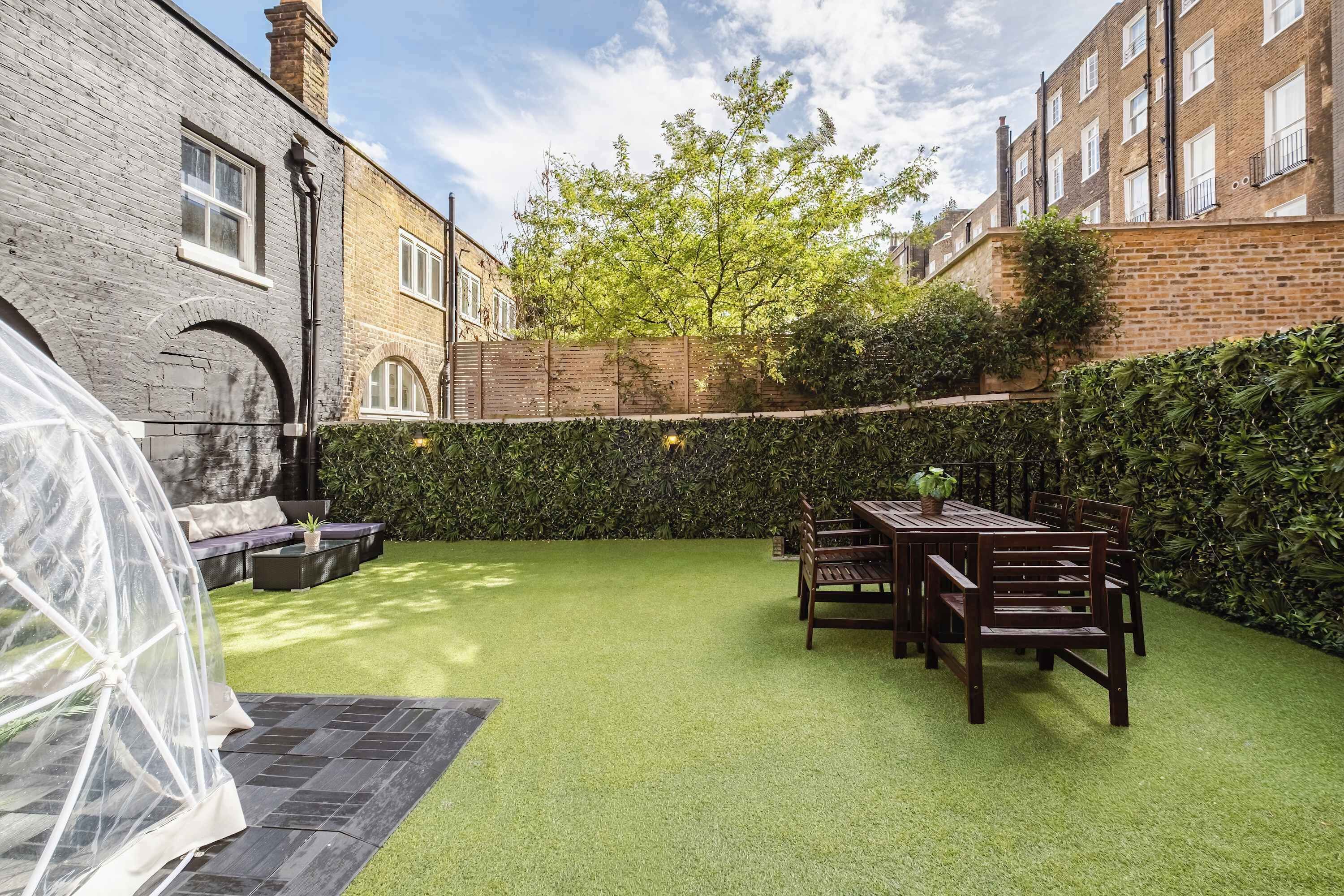 A courtyard with artificial grass, a wooden table and chairs, and a hedge.