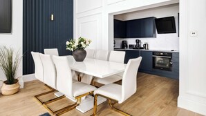 A modern dining area with a white table and chairs, a vase of flowers, and a kitchen in the background.