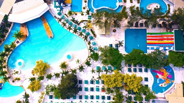 Vista aérea de un resort con múltiples piscinas, palmeras y tumbonas.