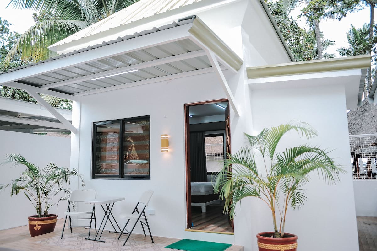 A modern, white building with a covered patio area, a bed, and potted plants.