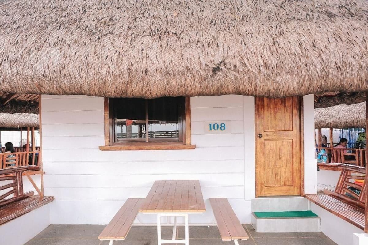 A thatched-roof building with a wooden door marked '108', a window, and a wooden table and bench in the foreground.