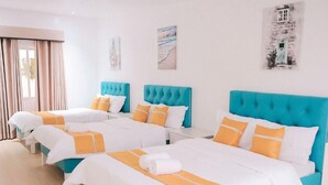 Habitación de hotel con tres camas, cada una con cabecero azul y ropa de cama blanca con detalles naranja. Hay dos cuadros en la pared, uno que representa una escena de playa y el otro, un edificio.