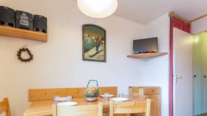 Une table à manger en bois, quatre chaises, un téléviseur fixé sur une étagère et un tableau encadré au mur.