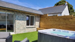 A backyard with a hot tub, wicker furniture, and a stone and wood exterior. - Maison Familiale Avec SPA Proche mer et Commodités (Trélévern)