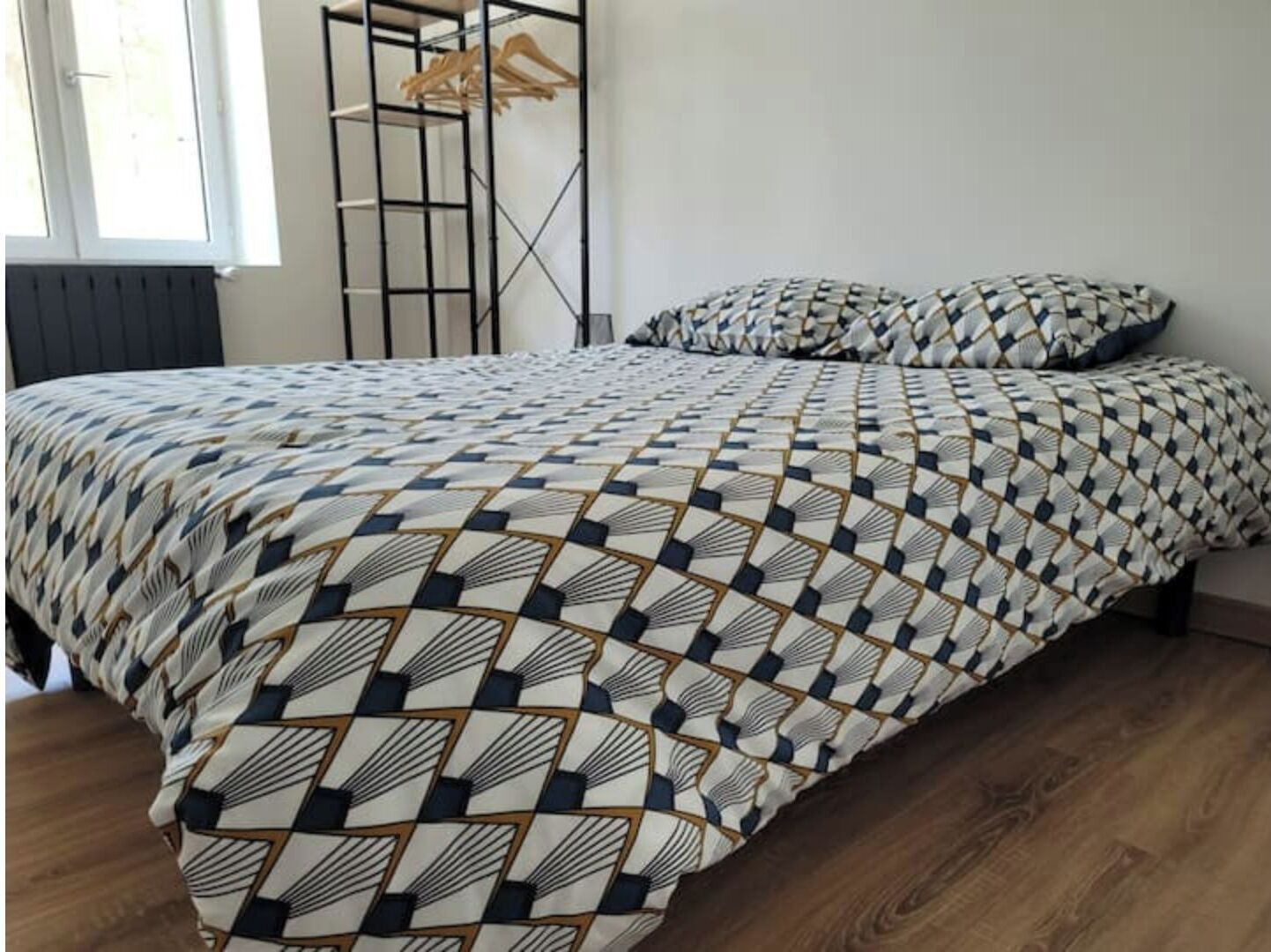 A bed with a geometric patterned bedspread and matching pillows, a wooden floor, a radiator, and a window with a view of the outside.