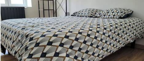A bed with a geometric patterned bedspread and matching pillows, a wooden floor, a radiator, and a window with a view of the outside.