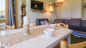 Ein gemütliches Zimmer mit Esstisch für zwei Personen, einer Couch, einem an der Wand befestigten Fernseher und einer Lampe.