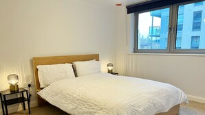 Una cama bien hecha con ropa blanca, dos lámparas de noche y una ventana con vistas a edificios.