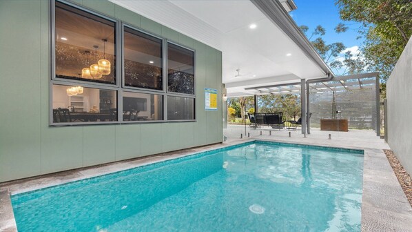 A swimming pool with a covered patio area, a dining area visible through large windows, and a pool safety sign. - EUCALYPTS - THE VINTAGE HOLIDAY HOUSES (Rothbury)