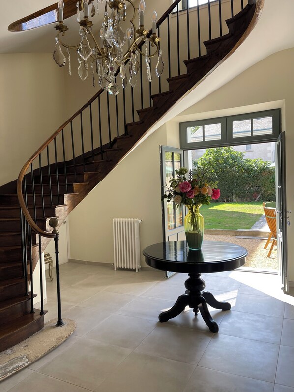 A staircase with a chandelier, a round table with a vase of flowers, and a view of a garden through an open door.
