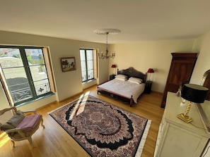A bedroom with a bed, two bedside lamps, a chandelier, a large window, a chair, and a rug.