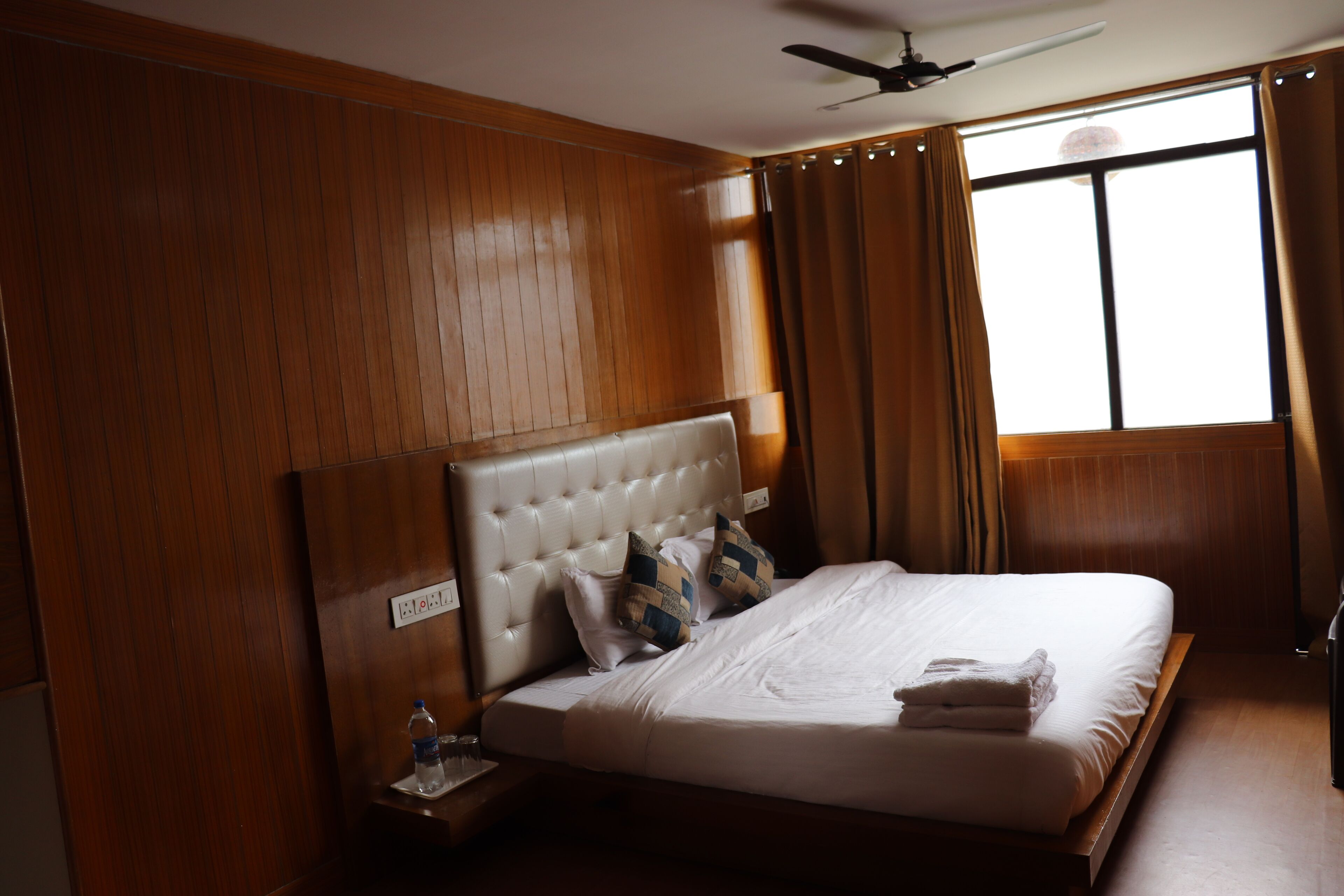 A hotel room with a wooden panel wall, a tufted headboard, a bed with white linens, two pillows, a folded towel, a bedside table with a water bottle, and a window with curtains.