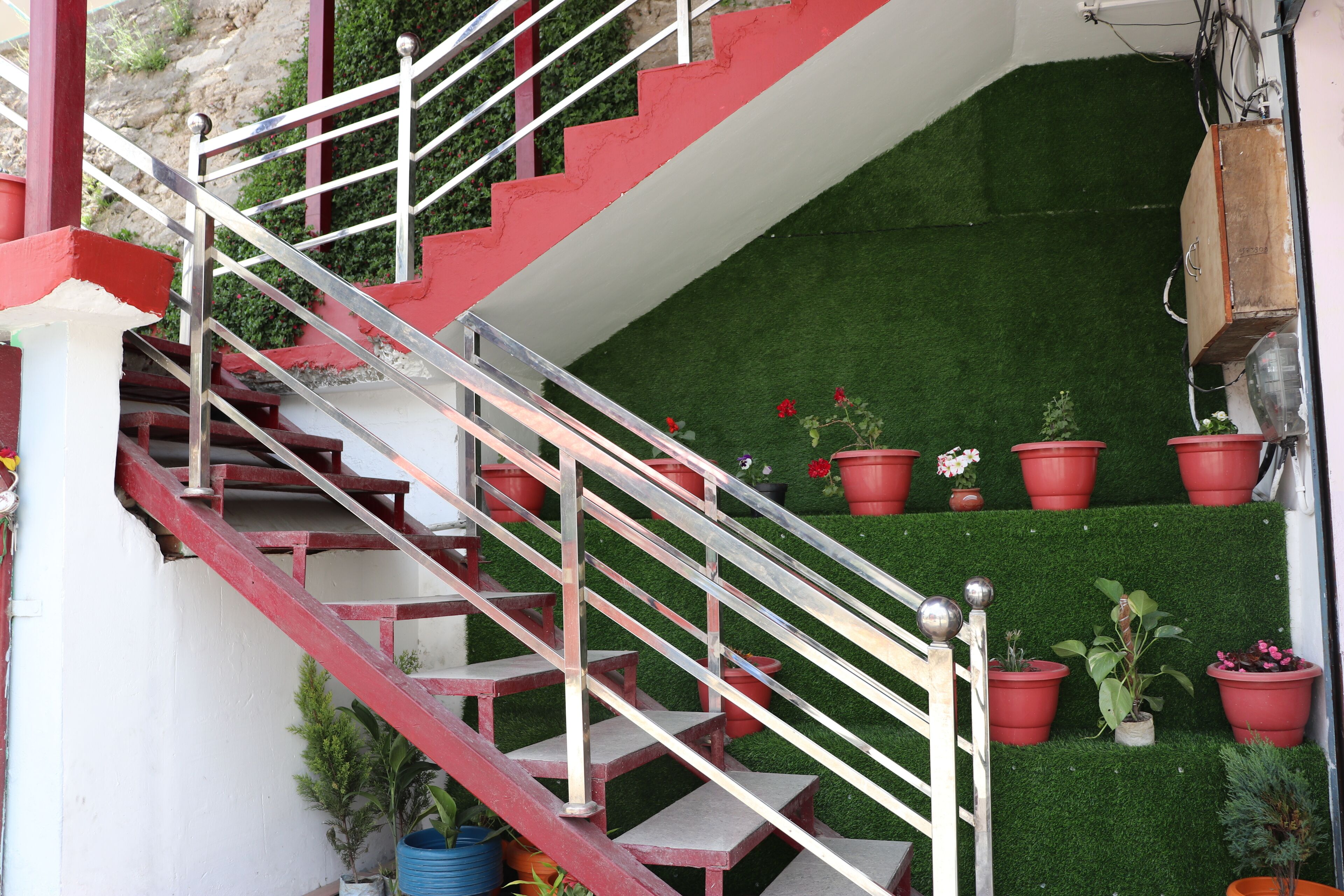 Un escalier avec des marches rouges et des rampes blanches, menant à une petite plateforme recouverte de gazon synthétique vert, agrémentée de plantes en pot.