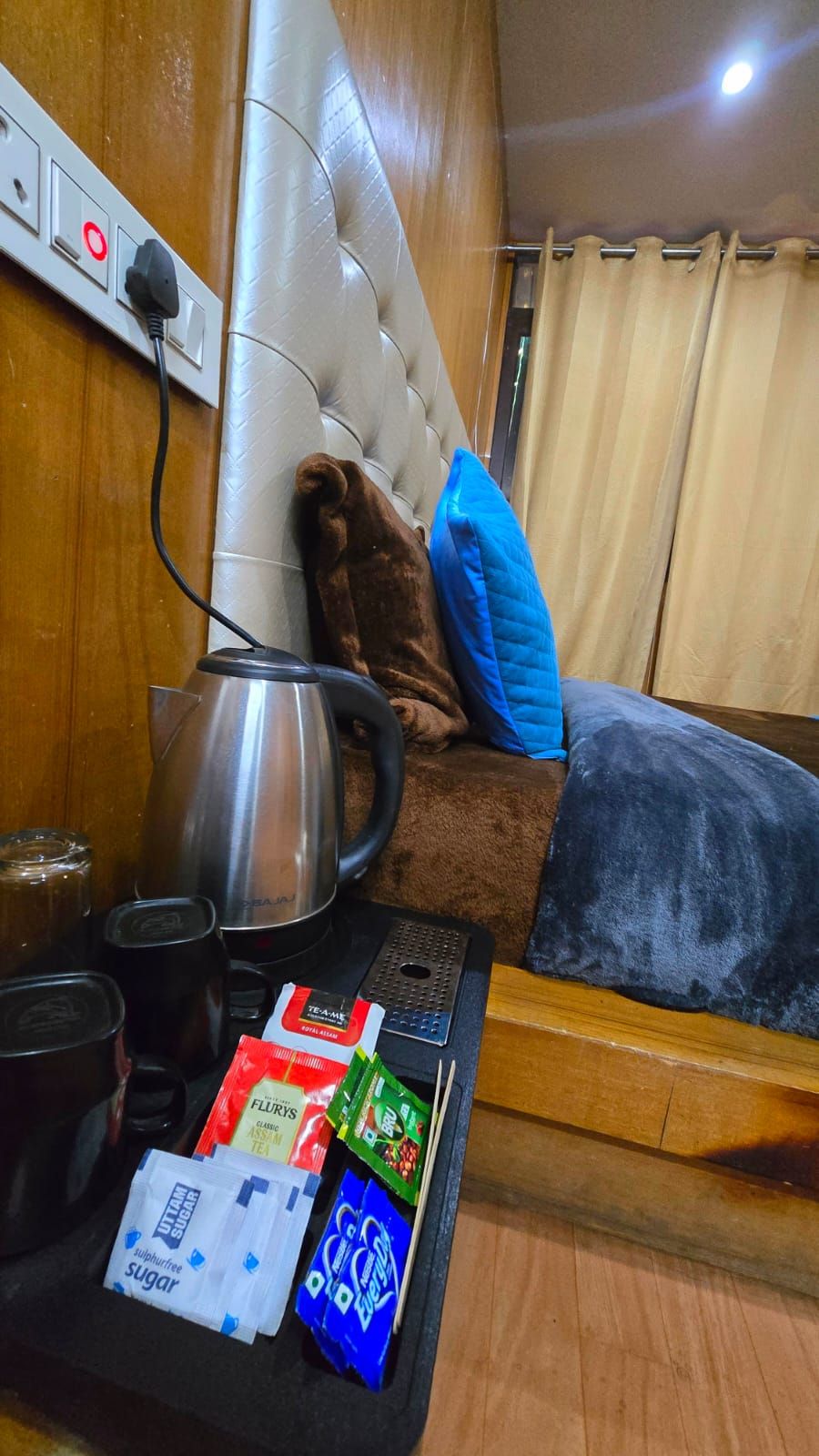 A cozy hotel room with a bed, a bedside table with a kettle, tea packets, and a remote control.