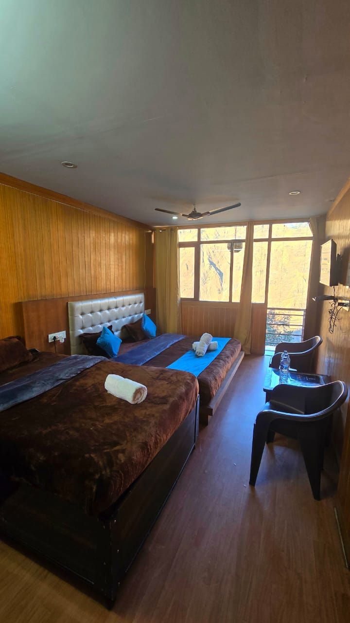 Quarto de hotel com duas camas, um ventilador de teto, janela com vista e uma pequena mesa com uma cadeira.