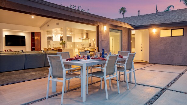 An outdoor dining area with a table and chairs, a sofa, and a kitchen visible through an open doorway. - 2 Modern Villas On the Golf Course | Putting Green, Pool, Hot Tub | Sleeps 22 (La Quinta)