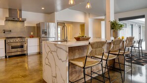 A modern kitchen with a marble island, stainless steel appliances, and a seating area with woven chairs. - 2 Modern Villas On the Golf Course | Putting Green, Pool, Hot Tub | Sleeps 22 (La Quinta)