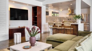 A modern living room with a sofa, a coffee table, and a TV. There is a kitchen area with a dining table and chairs. - 2 Modern Villas On the Golf Course | Putting Green, Pool, Hot Tub | Sleeps 22 (La Quinta)