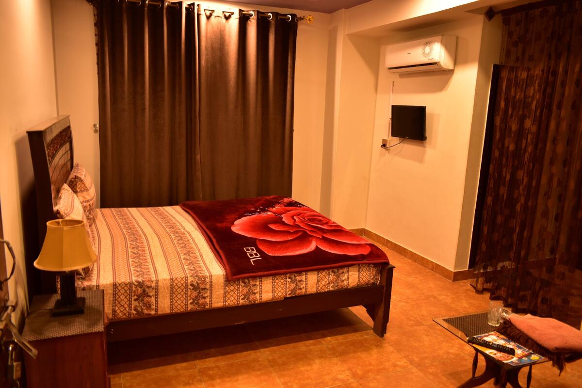 A bedroom with a bed, a bedside table with a lamp, a TV mounted on the wall, and an air conditioning unit.