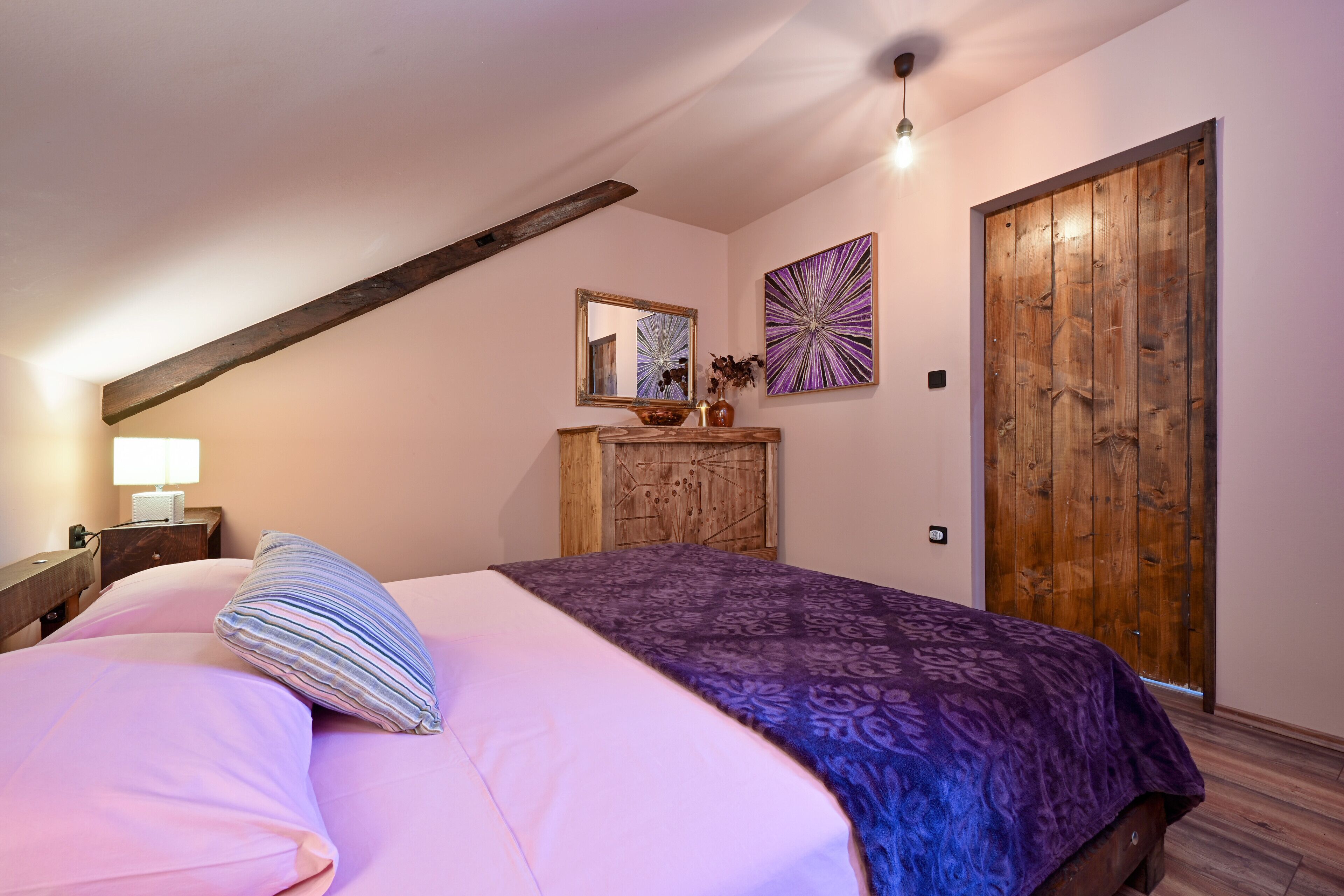 A bedroom with a sloped ceiling, a wooden wardrobe, a bed with pink and purple bedding, a nightstand with a lamp, and a mirror above the dresser.