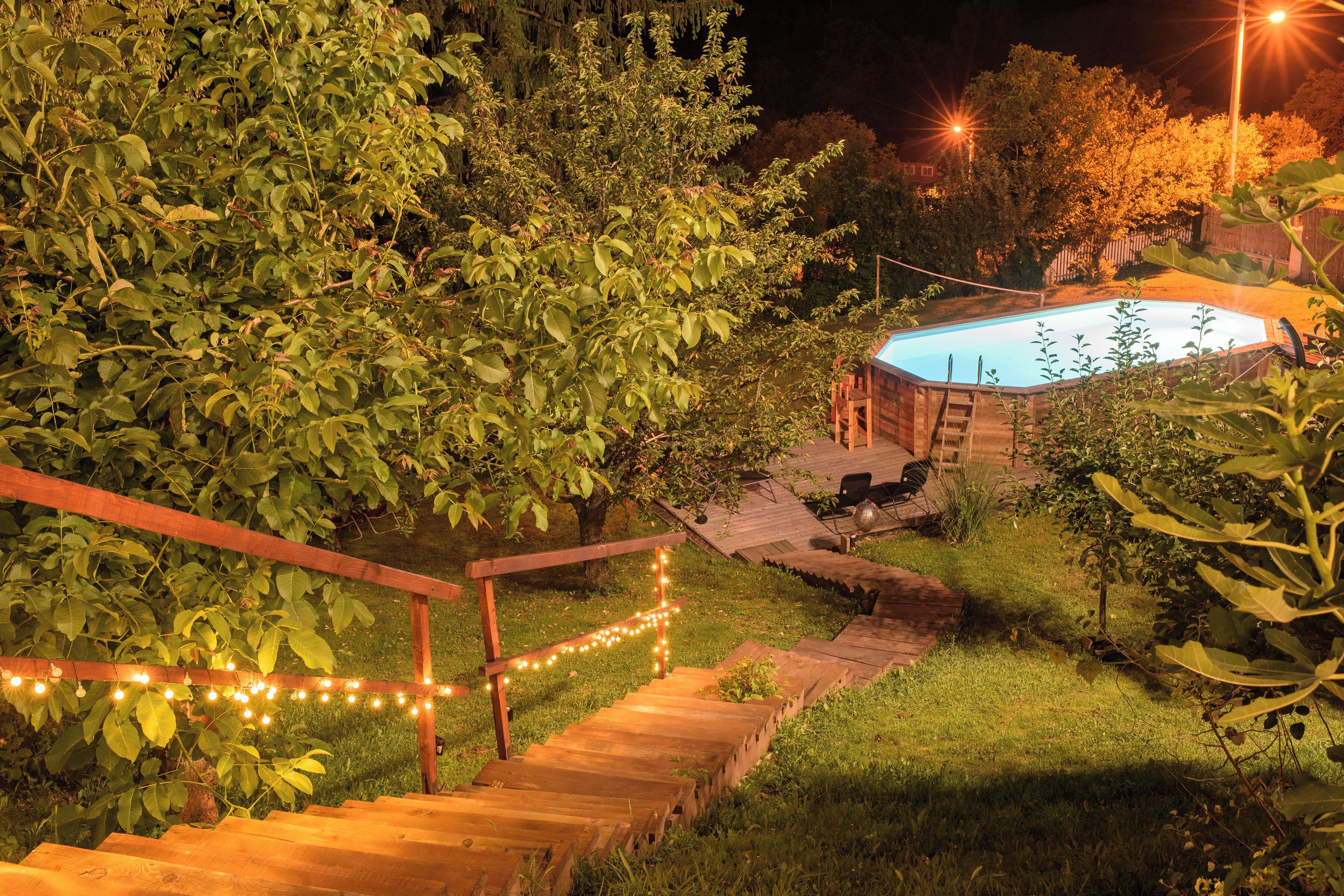Una escalera de madera que conduce a un área de piscina rodeada de vegetación, con luces colgantes a lo largo de los peldaños.