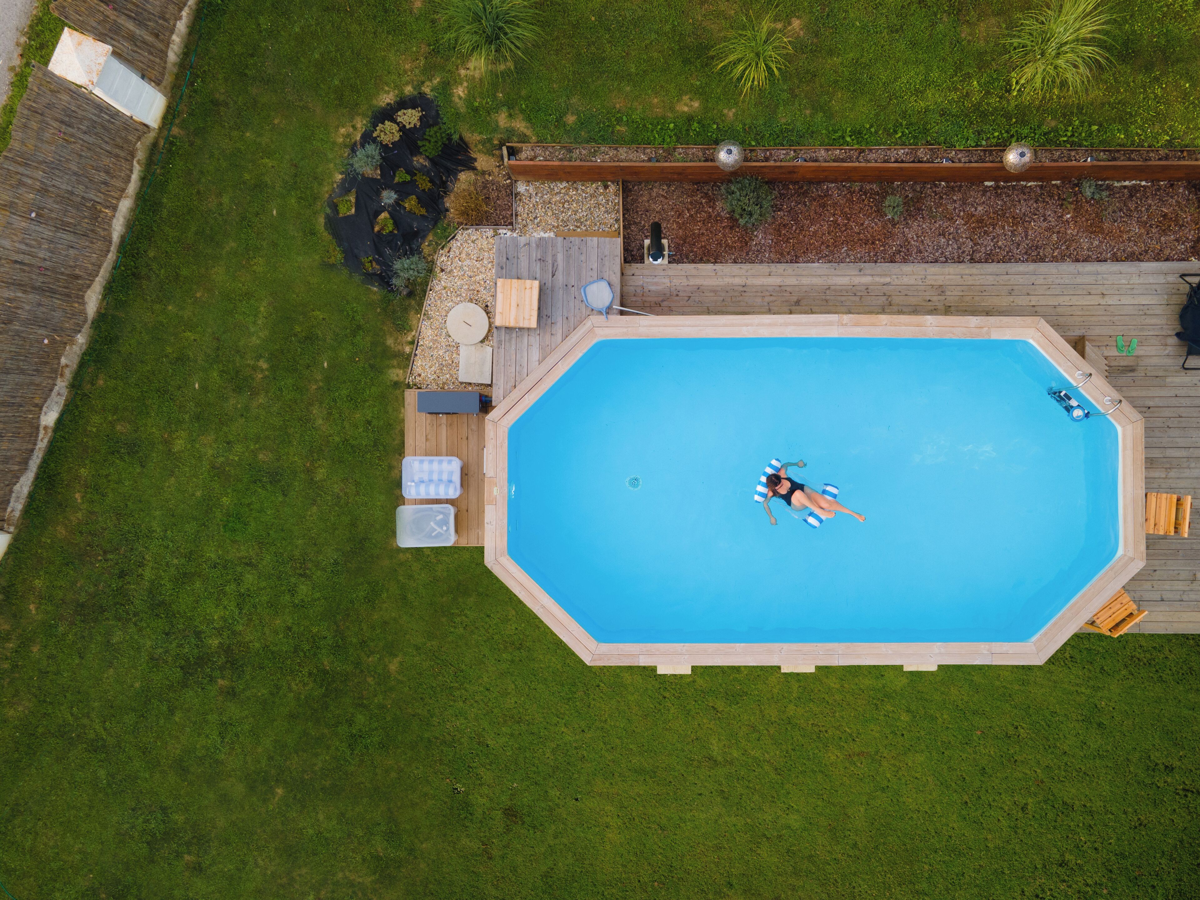 An octagonal above-ground pool with a wooden deck surrounding it, a small seating area, and a garden with plants and a bird bath.
