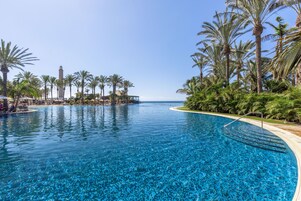 A large, clear blue pool with palm trees and a distant view of a lighthouse.