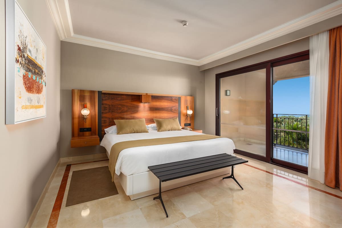 Une chambre d’hôtel avec un grand lit, une banquette, une tête de lit en bois, une table de chevet, une œuvre d’art accrochée au mur et un balcon avec vue.