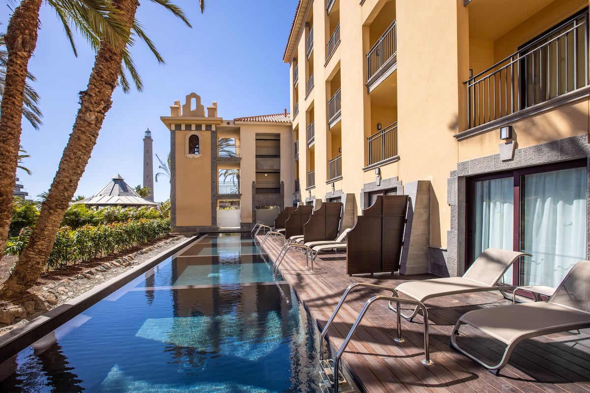 Un espace piscine avec des chaises longues, un palmier et un bâtiment doté de balcons.