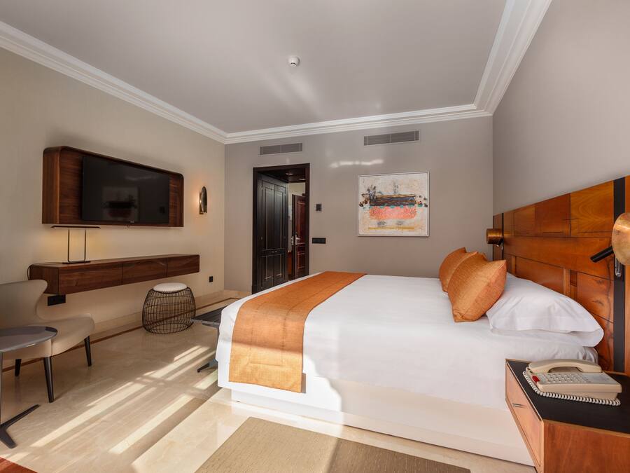 A hotel room with a large bed, a desk with a lamp, a television mounted on the wall, and a painting on the wall.