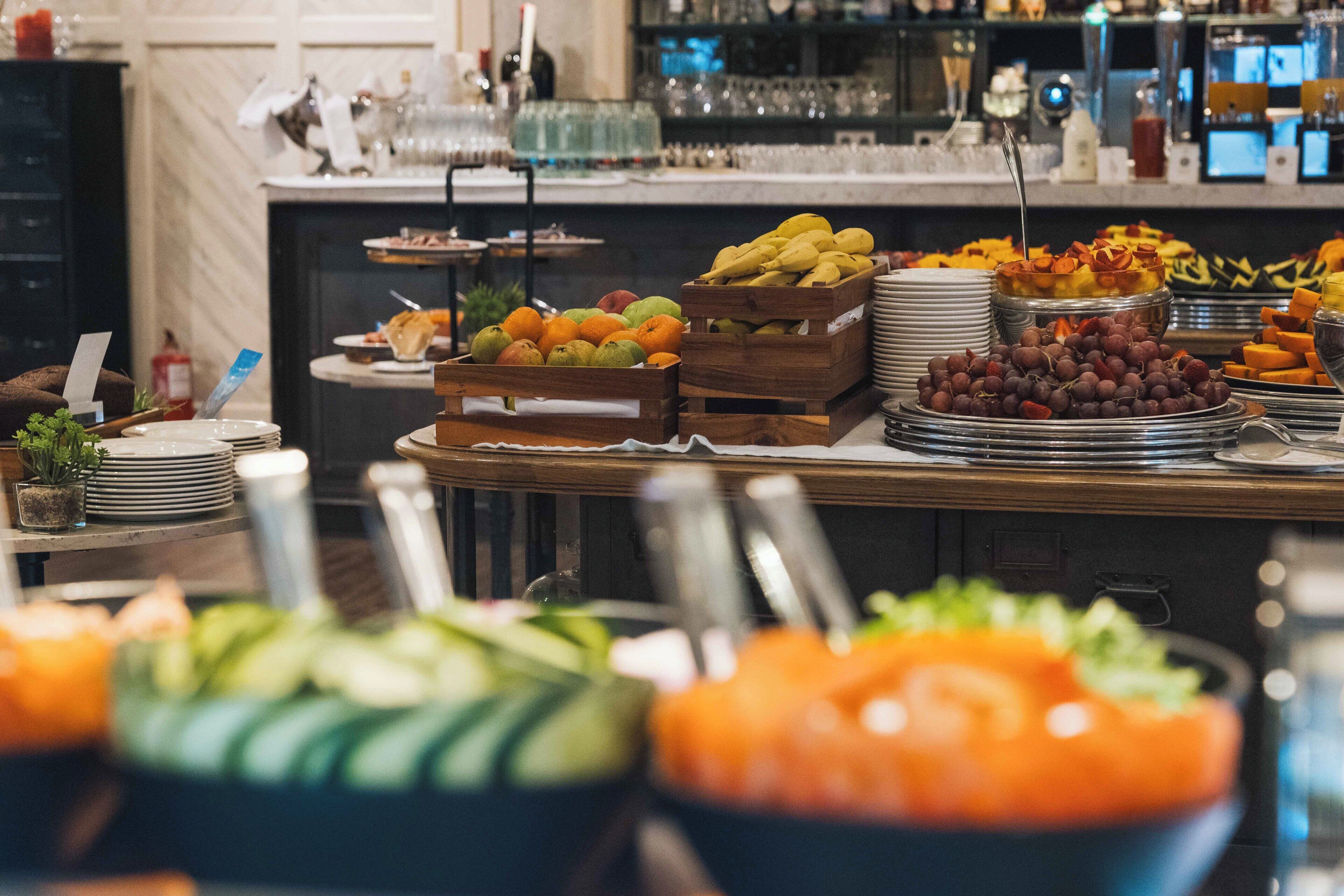 Un bufé con diversas frutas y verduras, una zona de bar con vasos y botellas, y un mostrador con platos y cubiertos.