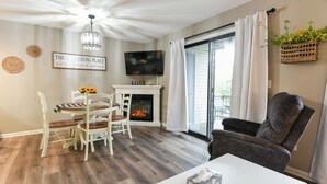 A living room with a fireplace, a dining table with chairs, a television, and a large window with curtains.