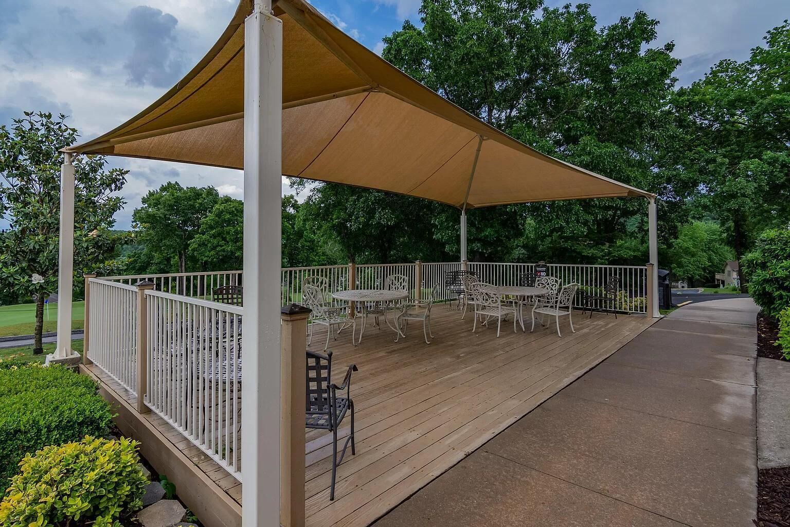 A covered outdoor patio with metal chairs and tables, surrounded by a white fence, set in a lush green environment.