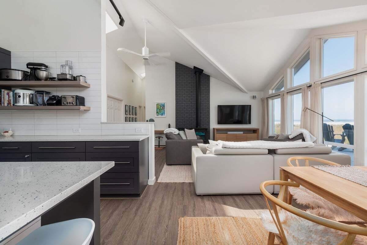 A modern kitchen with dark cabinets and white countertops, a dining area with a wooden table and chairs, and a living area with a sofa and a TV.