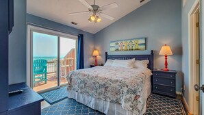 A bedroom with a bed, two bedside tables with lamps, a ceiling fan, a painting of beach huts, and a balcony with chairs. - Sunny Turtle: Turtle-tastic Oceanfront Stay! (Surf City)