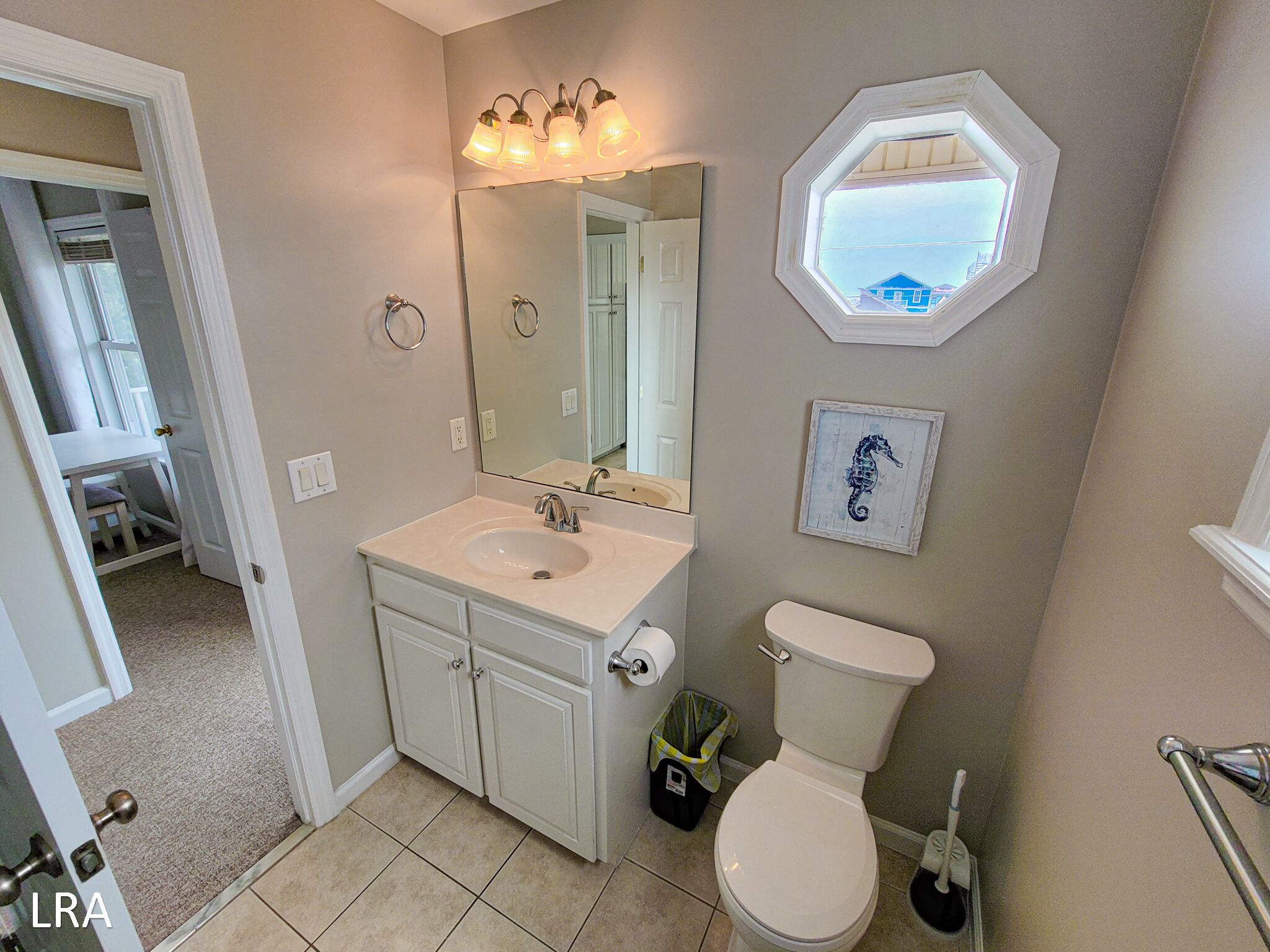 Une salle de bain avec un lavabo blanc, des toilettes et un miroir. Il y a une fenêtre donnant sur une maison à l’extérieur.