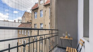 Een balkon met uitzicht op omliggende gebouwen, een klein tafeltje met een fles en glazen, en een houten stoel.