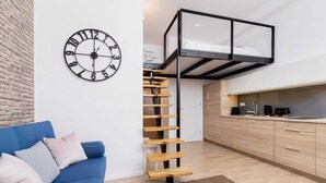 Un espace de vie moderne avec un canapé bleu, une échelle en bois, un coin cuisine et une horloge murale.