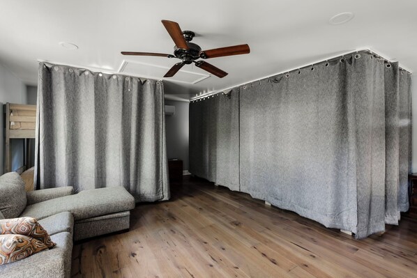 A room with a ceiling fan, grey curtains, a wooden floor, and a grey sofa with a patterned cushion.