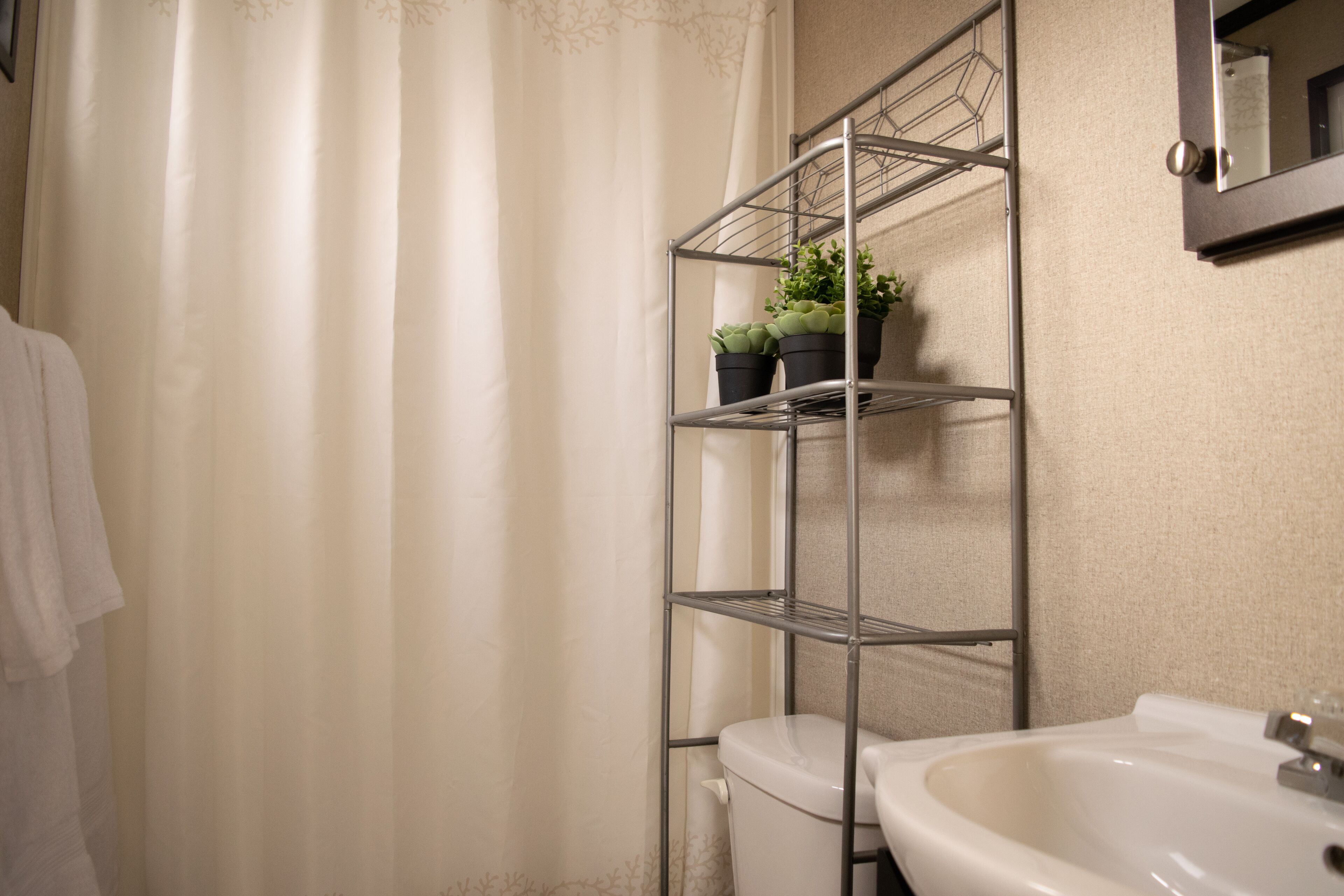 Un baño con lavamanos blanco, inodoro, estante metálico con plantas en macetas y cortina de ducha.