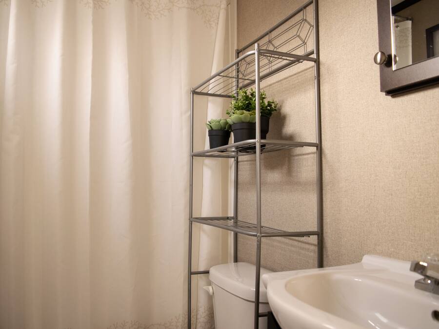 Un baño con lavamanos blanco, inodoro, estante metálico con plantas en macetas y cortina de ducha.