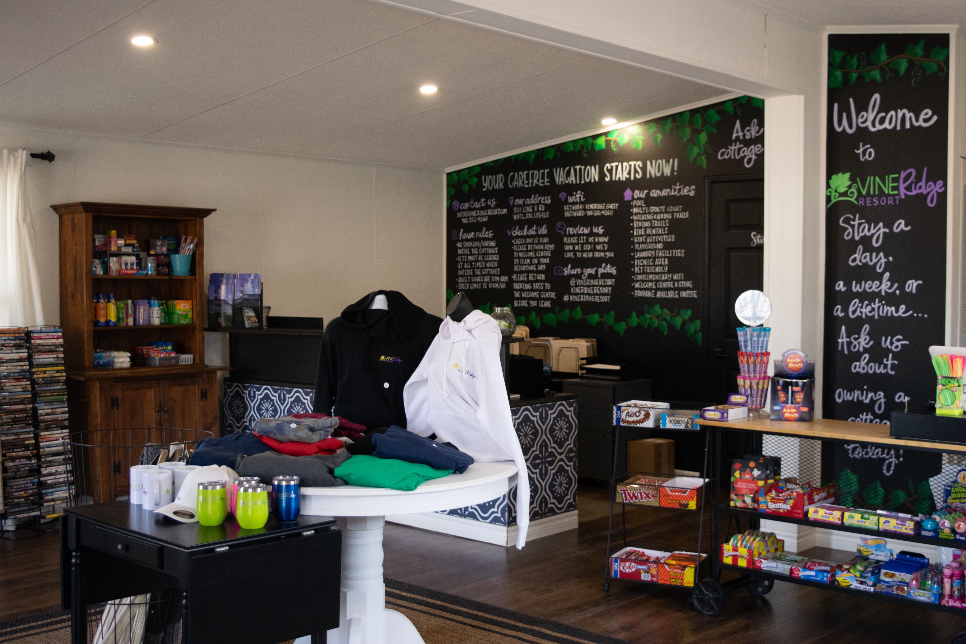 A room with a bed, a table with drinks, a shelf with snacks, and a chalkboard with information.