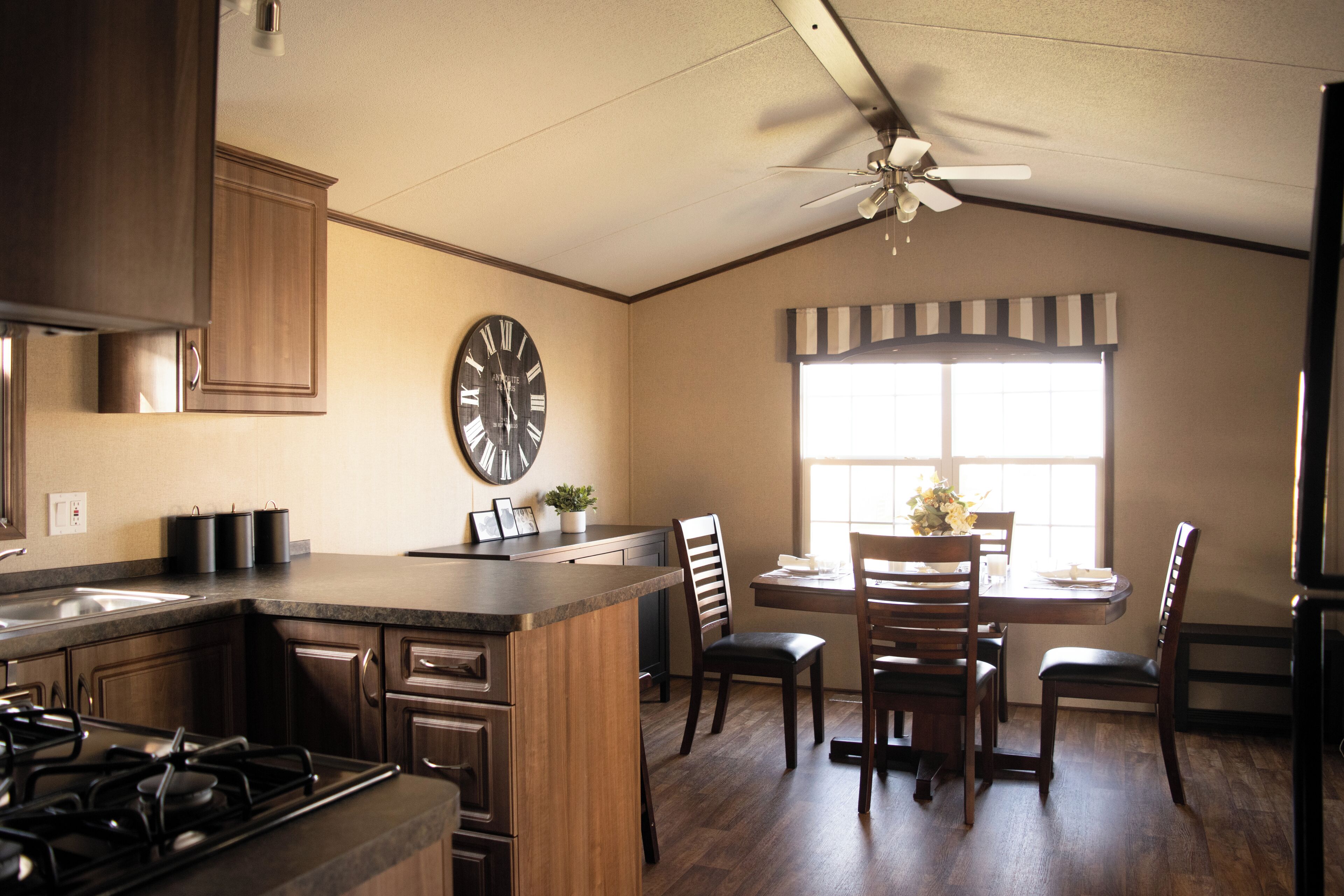 A kitchen with wooden cabinets, a stove, a dining table with chairs, and a large clock on the wall.